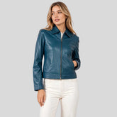 A woman modeling a teal blue faux leather jacket with a front zipper and collar, paired with white pants, against a solid gray background.