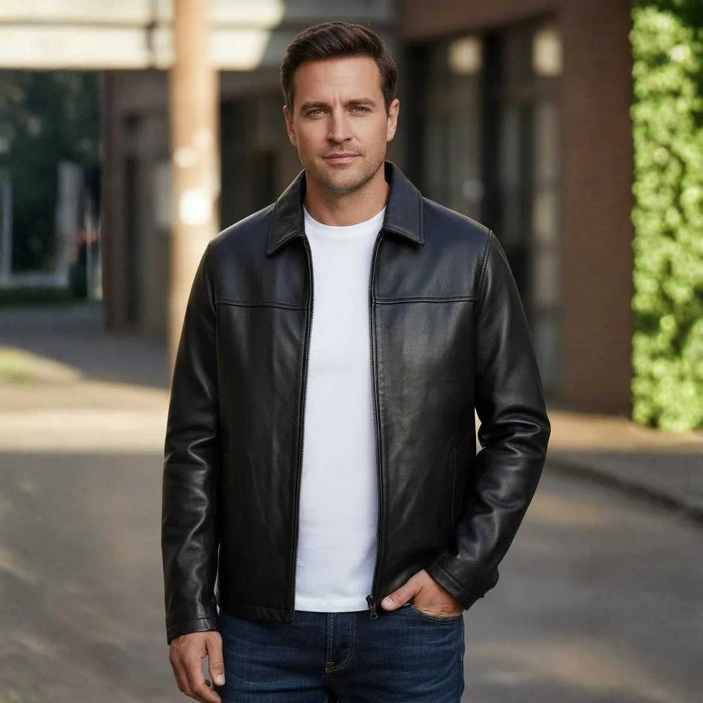 A man wearing a black leather jacket over a white t-shirt and blue jeans, standing outdoors on a paved walkway with a blurred urban background.