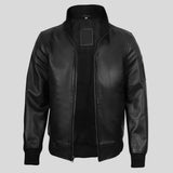A black leather bomber jacket with a stand-up collar, front zipper, ribbed cuffs and hem, and a rectangular patch on the chest, shown against a light gray background.