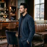 A man wearing a dark navy blue suede jacket and matching navy trousers stands in a dimly lit, upscale bar or lounge setting with brown leather seating and warm ambient lighting.