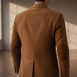 Back view of a man wearing a camel brown suit jacket with dark brown elbow patches, standing in a softly lit room with warm sunlight on the background.