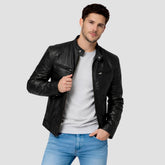 A man wearing a black leather jacket over a light gray t-shirt and blue jeans, standing against a solid light gray background.