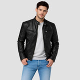 A man wearing a black leather motorcycle jacket over a light gray crewneck sweatshirt and blue jeans, standing against a solid light gray background.