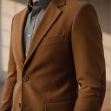 A close-up of a man wearing a brown tailored blazer over a dark gray button-up shirt, cropped from the neck to the waist against a softly lit indoor background.