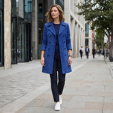 A woman walking on a city sidewalk wearing a bright blue double-breasted coat over a dark top and dark trousers, with white sneakers.