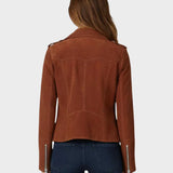 A woman viewed from the back wearing a cropped brown suede jacket with silver zipper details on the sleeves, paired with dark blue jeans, against a light gray background.