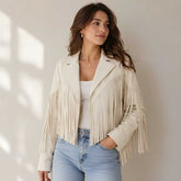 A woman wearing a cream-colored faux leather fringe jacket over a white tank top and light blue jeans, standing against a neutral beige wall with soft window shadows.