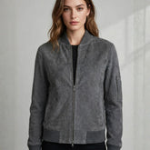 A woman wearing a gray suede bomber jacket with a front zipper, ribbed collar, cuffs, and hem, paired with a black top and black bottoms, set against a light gray textured wall.
