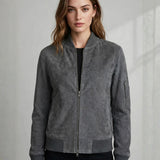 A woman wearing a gray suede bomber jacket with a front zipper, ribbed collar, cuffs, and hem, paired with a black top and black bottoms, set against a light gray textured wall.