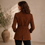A woman wearing a brown suede trench coat is shown from behind, standing in a warm, sunlit room next to a large rustic vase with dried branches.