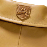 Close-up of a tan leather item featuring a stitched shield-shaped logo patch on the flap.