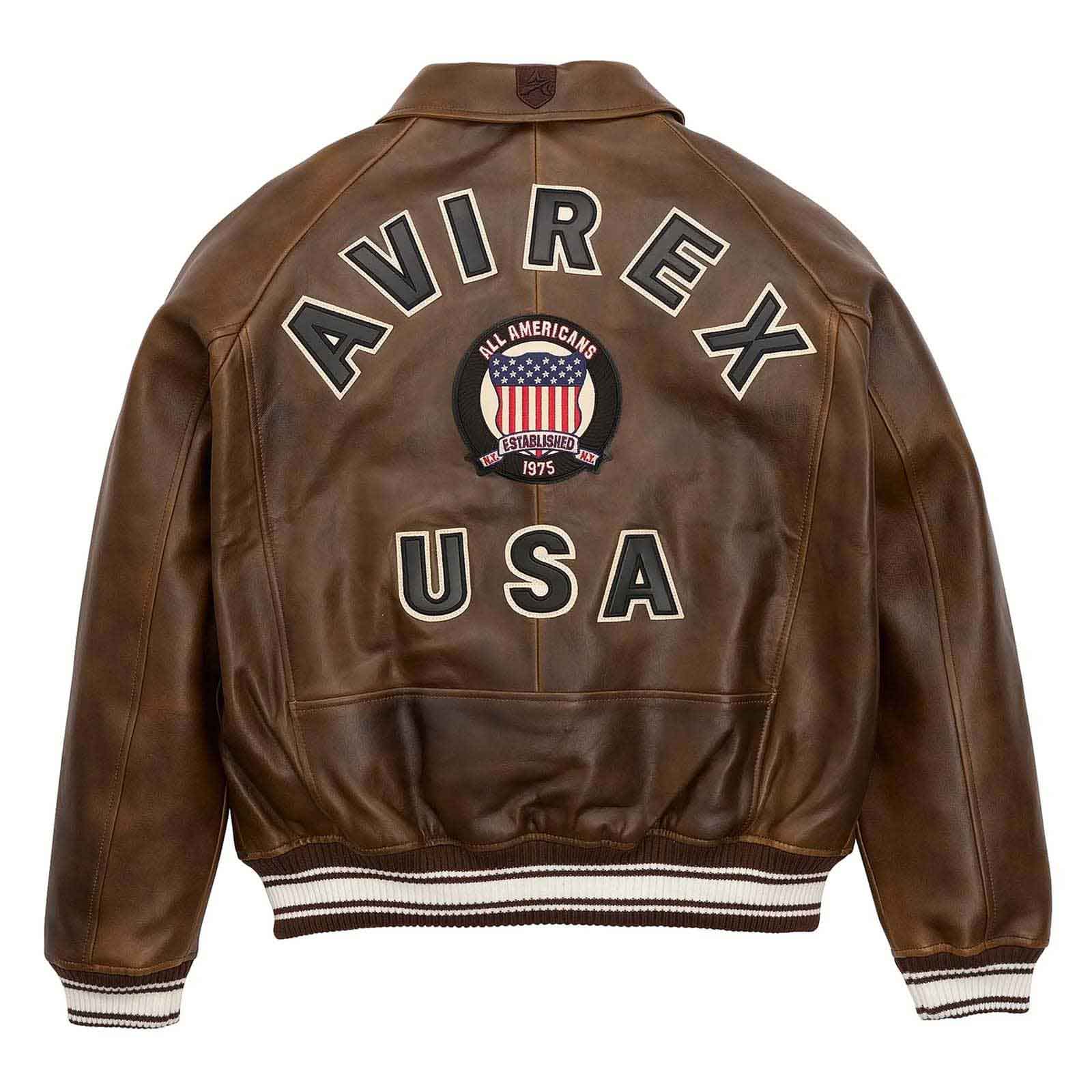 A brown leather jacket shown from the back, featuring 'AVIREX USA' embroidered in large letters and a circular 'All Americans 1975' patch in the center, with ribbed brown and white striped cuffs and hem.
