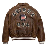 A brown leather jacket shown from the back, featuring 'AVIREX USA' embroidered in large letters and a circular 'All Americans 1975' patch in the center, with ribbed brown and white striped cuffs and hem.