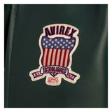 An embroidered patch featuring a shield design with red and white stripes, a blue field with white stars, and the text 'AVIREX' and 'ESTABLISHED 1975', shown on a dark green textured surface.