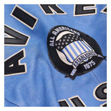 Close-up of a light blue sports jersey with large black and white lettering and a circular 'All Americans Established 1975' patch on the chest.
