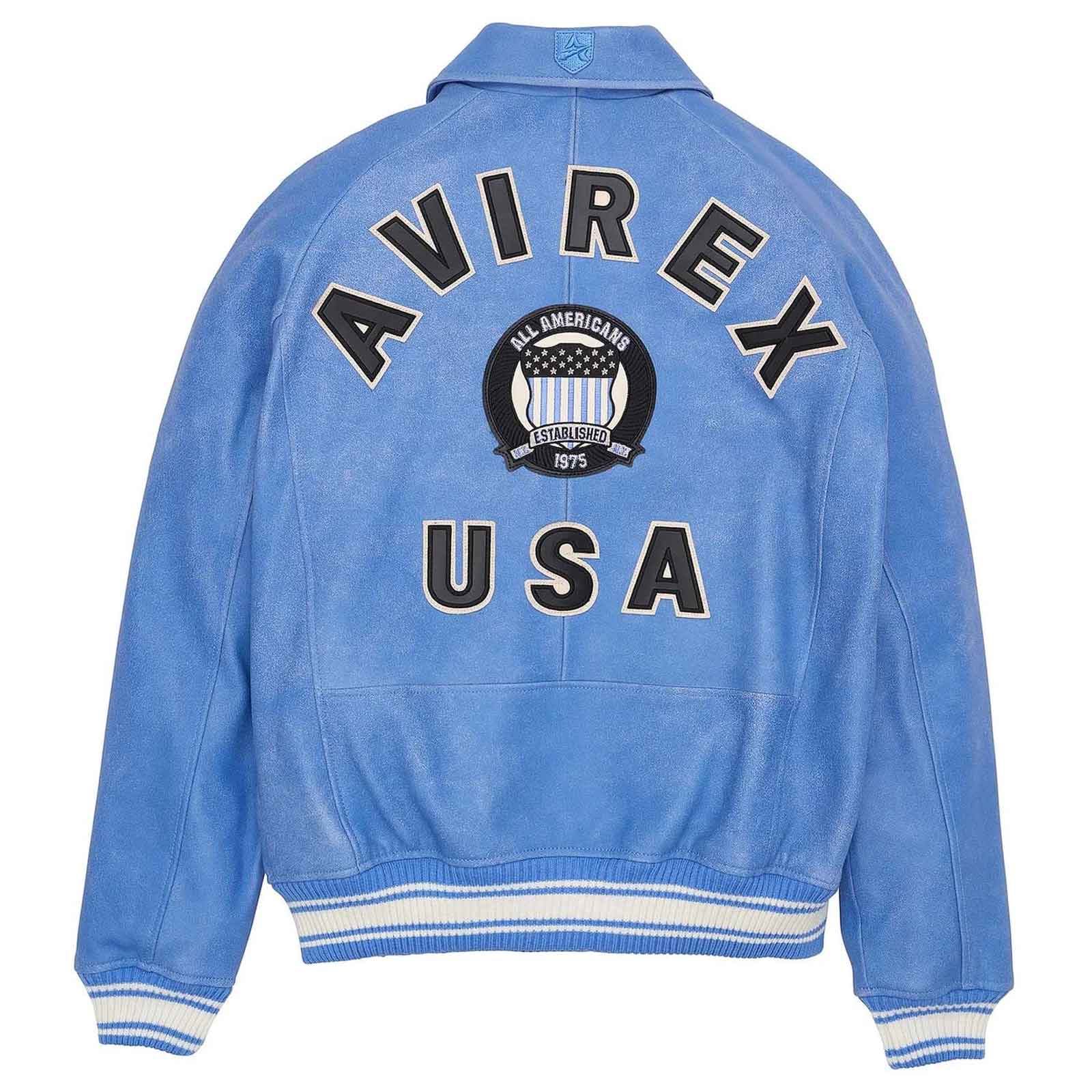 A light blue suede-like varsity jacket shown from the back, featuring 'AVIREX USA' in large black letters with white outlines and a circular 'All American' crest patch in the center. The jacket has white and blue striped ribbed cuffs and hem.