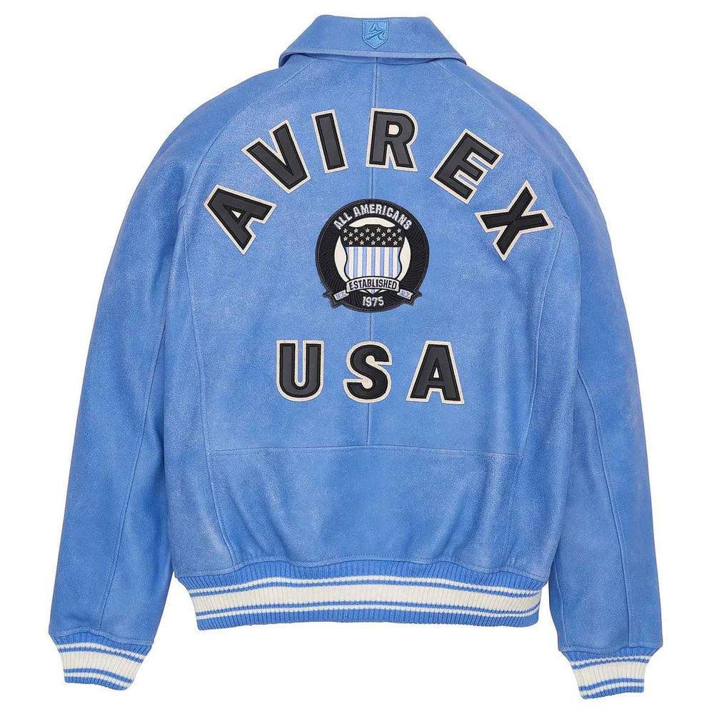 A light blue suede-like varsity jacket shown from the back, featuring 'AVIREX USA' in large black letters with white outlines and a circular 'All American' crest patch in the center. The jacket has white and blue striped ribbed cuffs and hem.