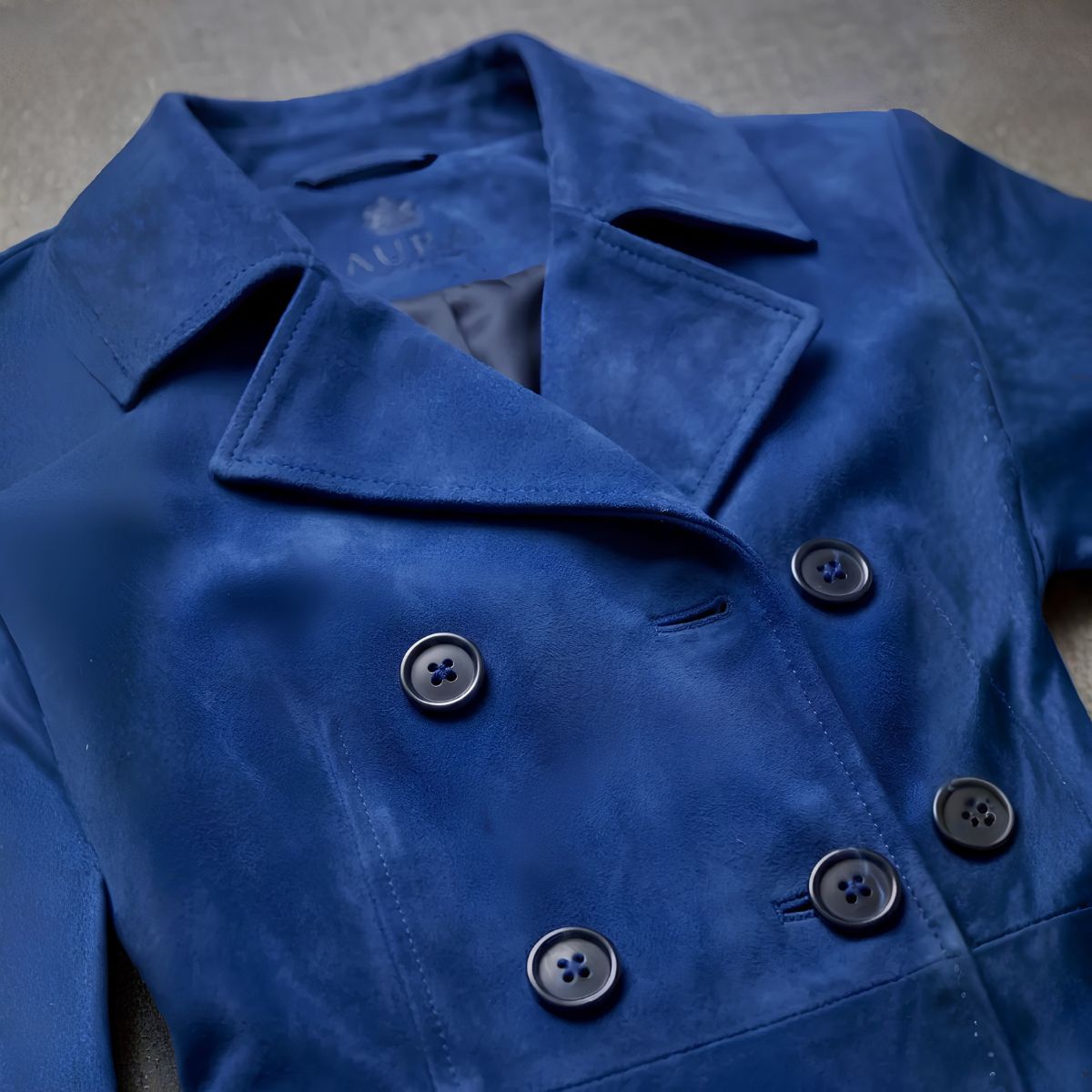 A close-up of a royal blue double-breasted coat or jacket with dark buttons, laid flat on a neutral textured surface. 