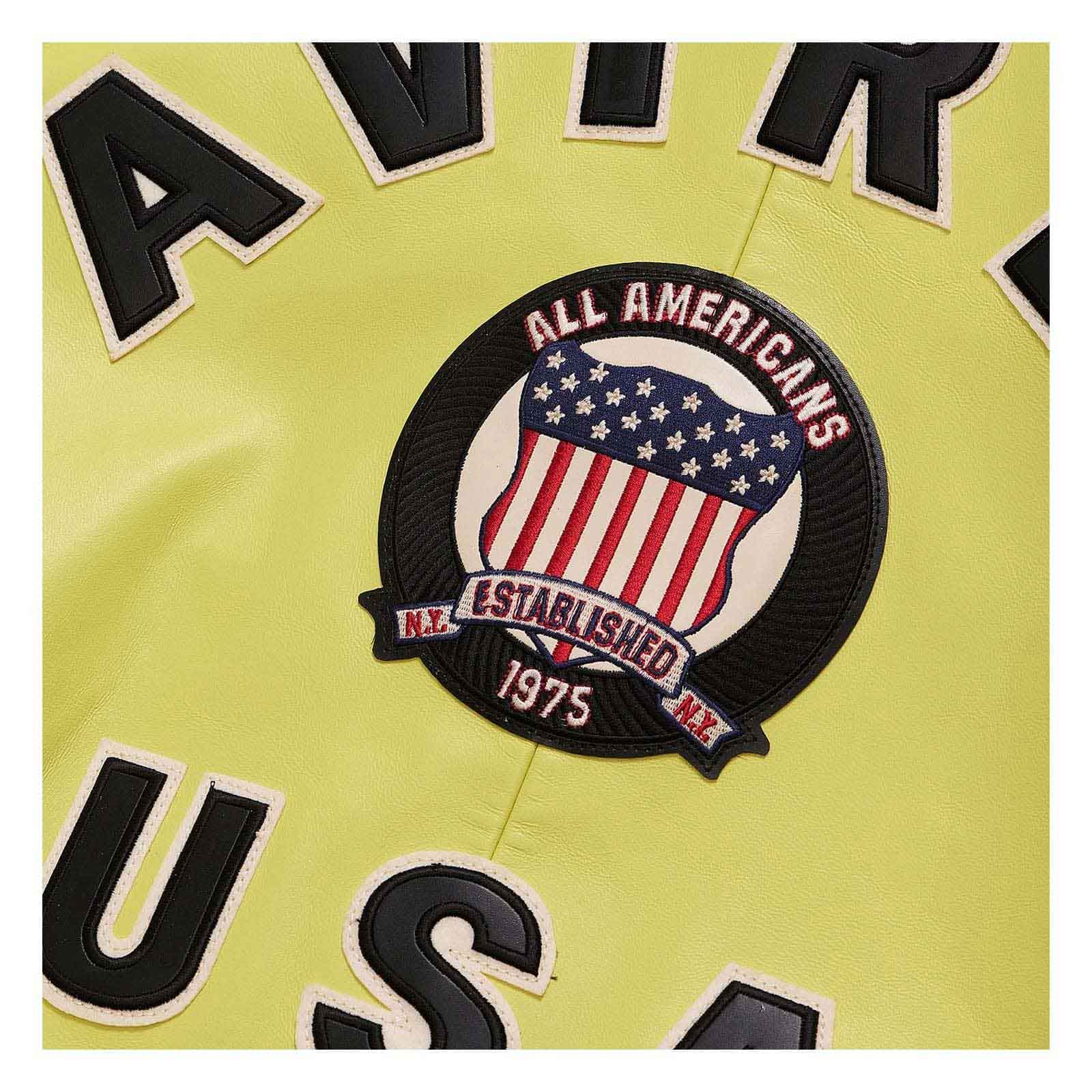 Close-up of a light yellow sports jersey with a circular 'All Americans Established 1975' patch featuring an American flag design, with black and white lettering partially visible.