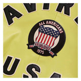 Close-up of a light yellow sports jersey with a circular 'All Americans Established 1975' patch featuring an American flag design, with black and white lettering partially visible.