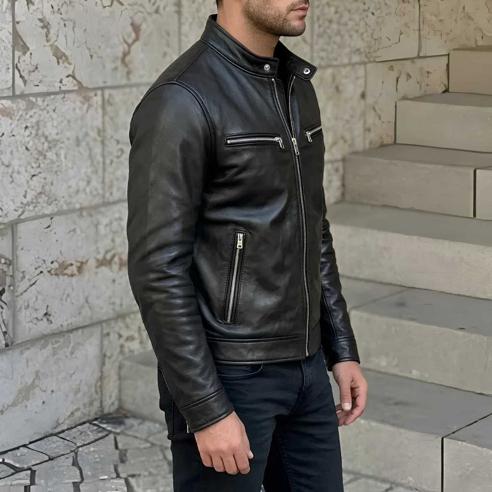 A man wearing a black leather motorcycle jacket with silver zippers, standing in front of a stone wall and steps.