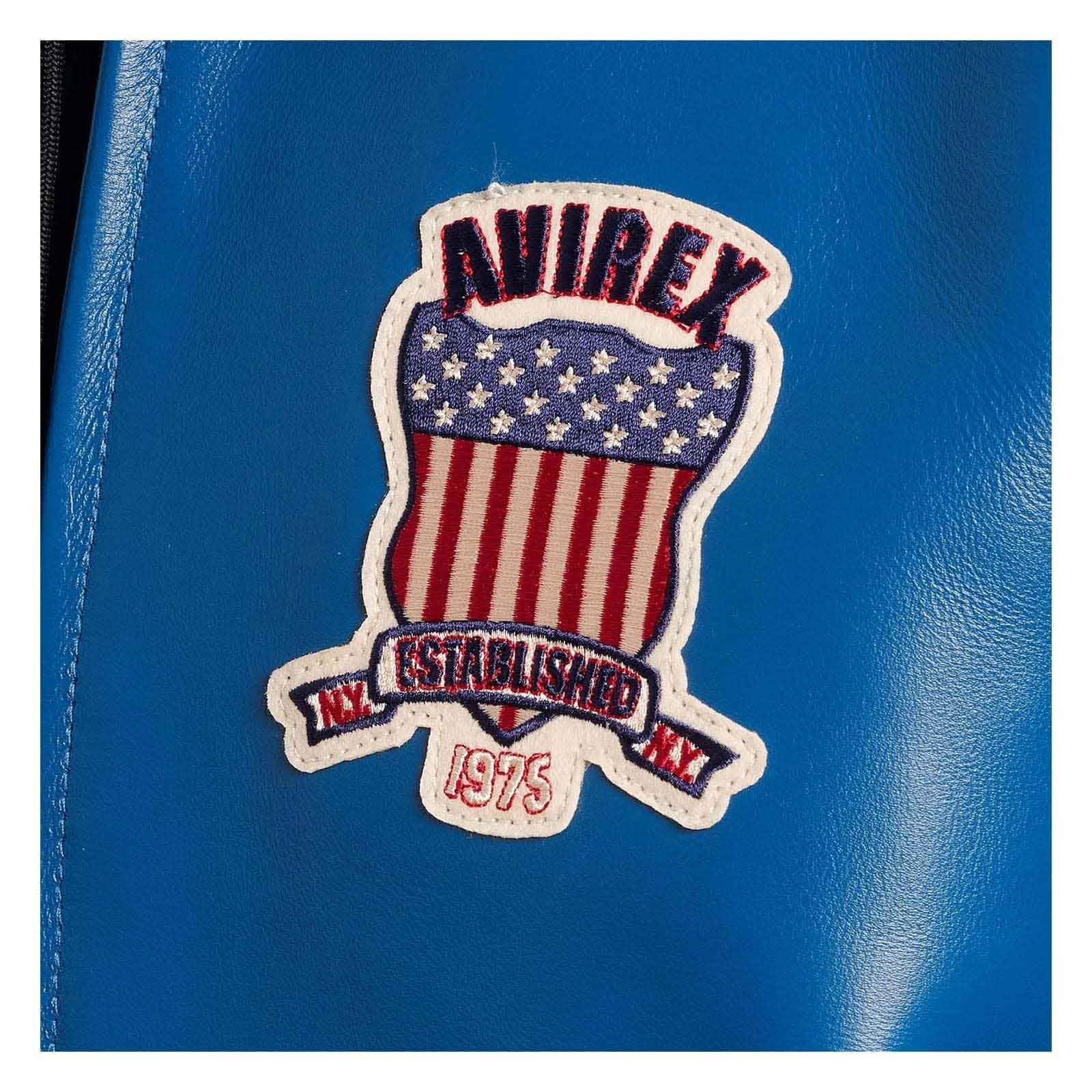 A close-up of a blue textured leather or faux-leather surface featuring an embroidered patch with the brand name 'AVIREX' above an American flag motif and 'ESTABLISHED 1975' on a banner.