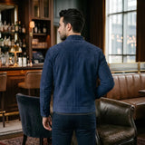 A man wearing a navy blue suede jacket and matching navy pants, standing in a dimly lit bar with brown leather seating and warm lighting.