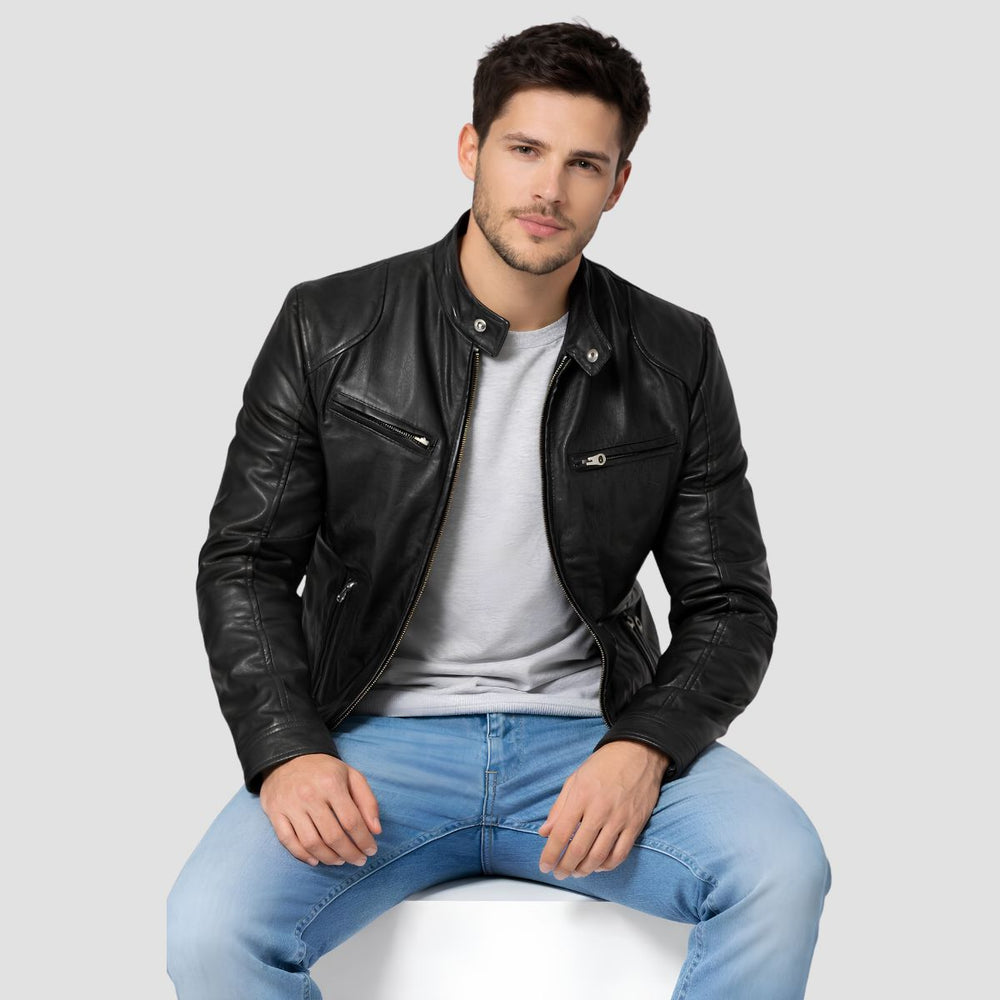 A man wearing a black leather moto jacket over a light gray t-shirt and blue jeans, sitting on a white stool against a light gray background.