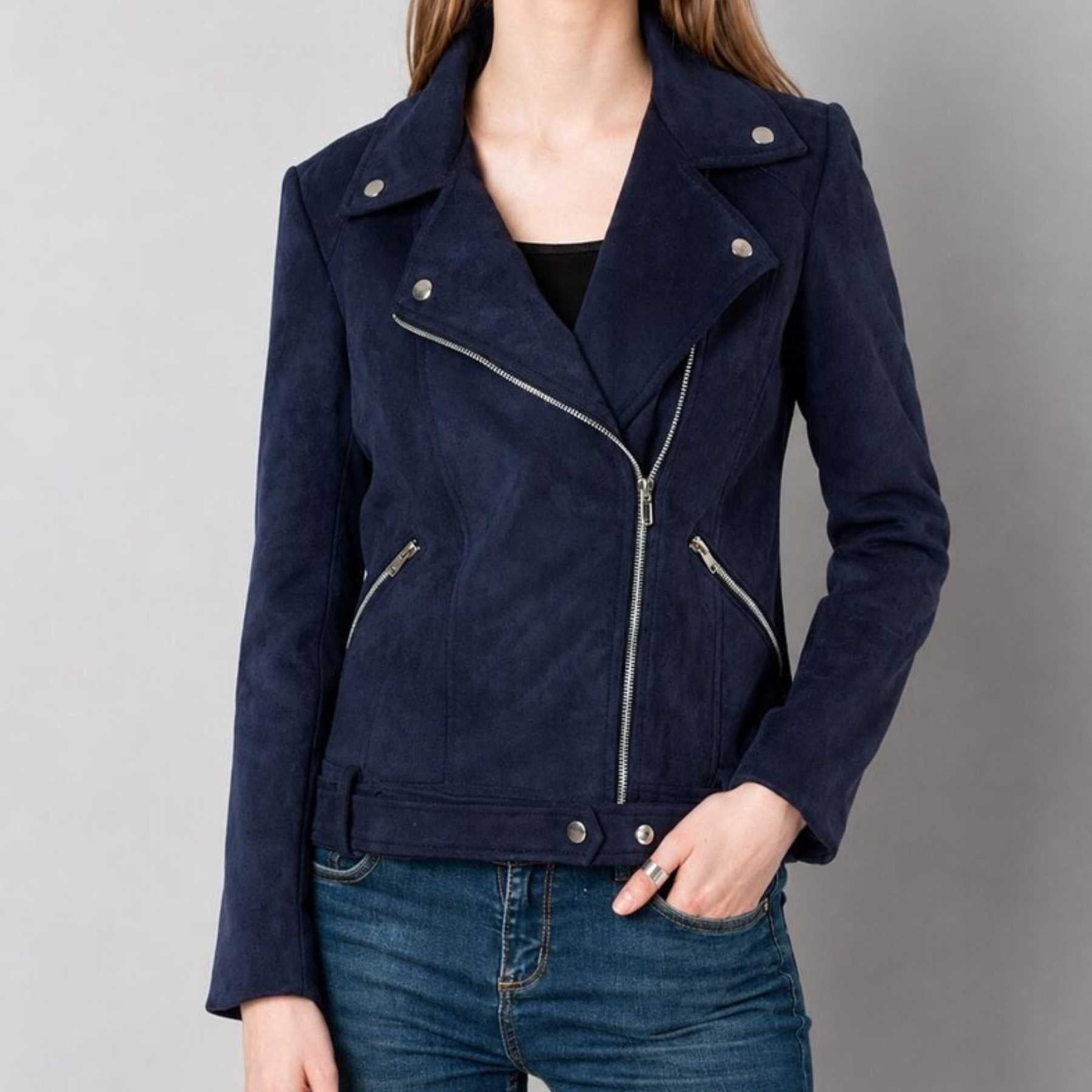 A woman wearing a navy blue suede moto jacket with silver zippers and snap buttons, paired with blue jeans, shown against a plain gray studio background.