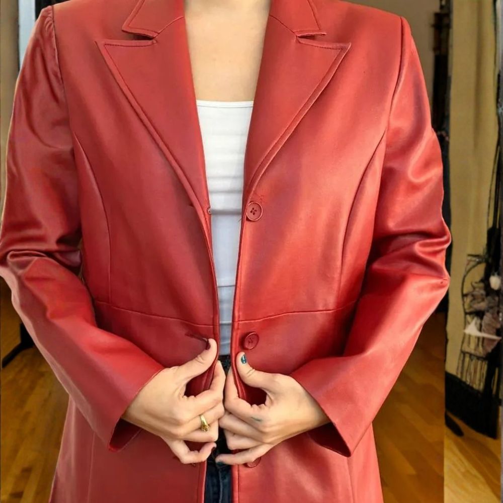 A person wearing a structured, long red faux-leather blazer over a white tank top, standing in an indoor setting with wooden floors.