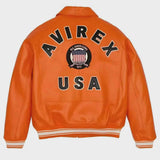 Bright orange leather jacket shown from the back, featuring large black varsity-style lettering reading 'AVIREX' with 'USA' below and a circular 'All American' crest patch in the center. The jacket has striped ribbed cuffs and hem and is photographed on a light gray background.