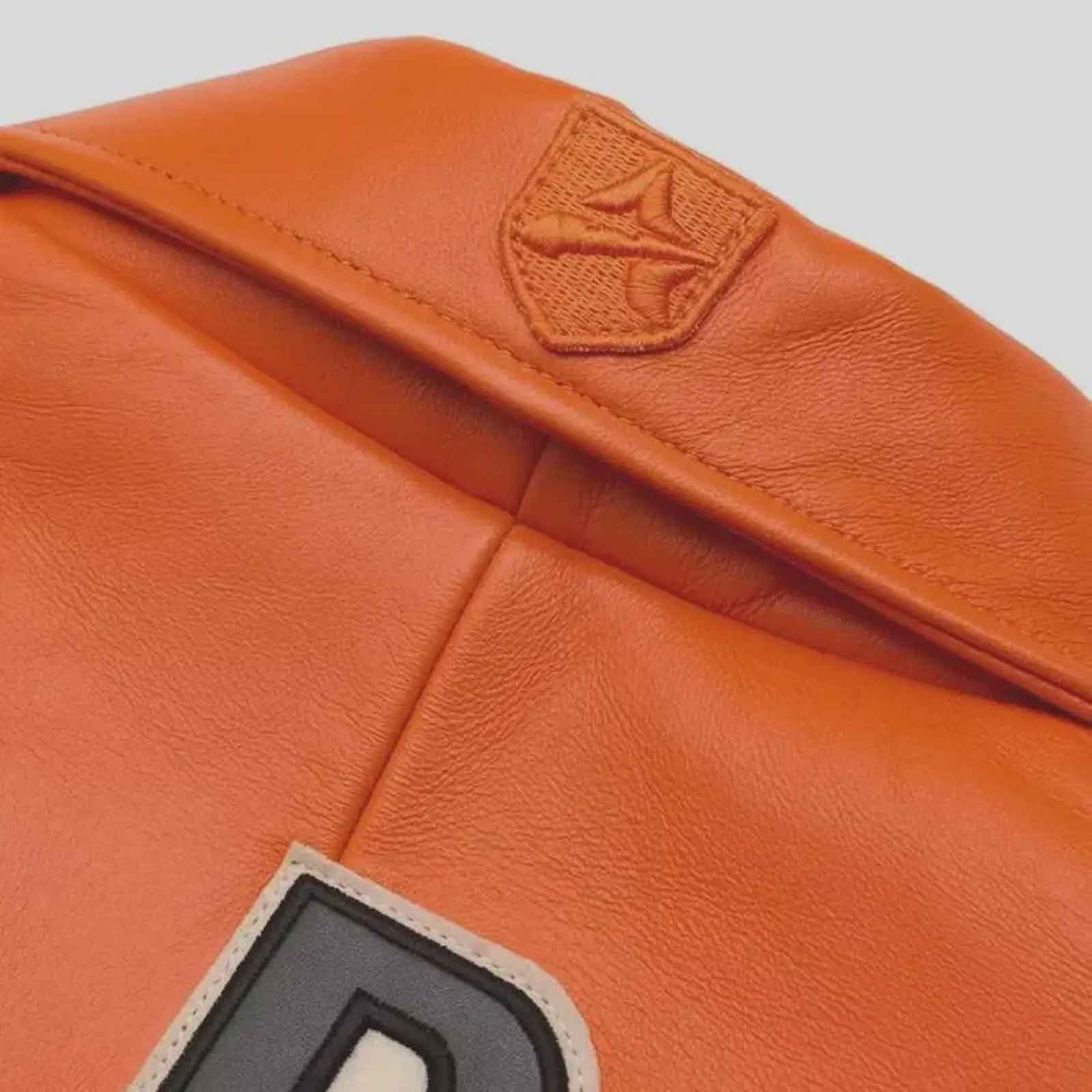 Close-up of an orange leather bag showing a stitched shield patch with a stylized bird logo and part of a large letter patch on the lower left.