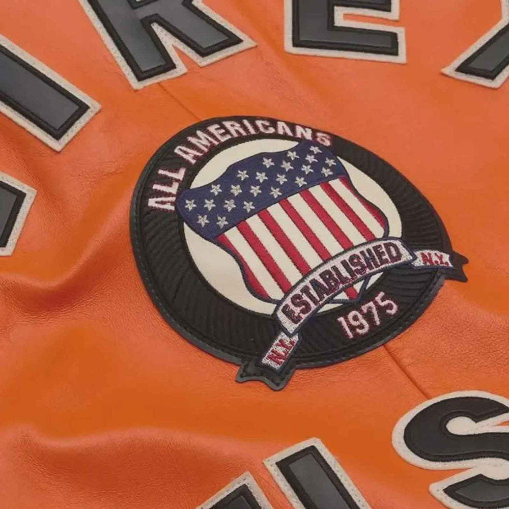 Close-up of an orange leather jacket featuring a circular 'All Americans Established 1975' patch with an American flag design, alongside partial black and white lettering.