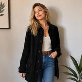 A woman wearing a black button-front coat over a white top and blue jeans, standing indoors against a light wall with framed wall art and a plant partially visible.