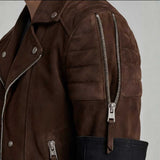 Close-up of a brown suede moto jacket with silver metal zippers, snap buttons, and a dark navy fabric panel at the hem, shown on a model against a light gray background.