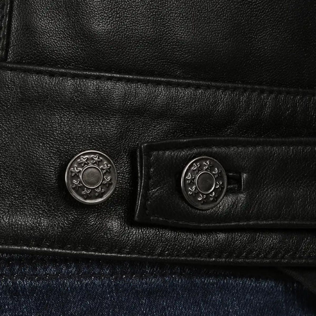 Close-up of a black leather jacket detail showing two round silver-tone decorative buttons on a strap, with dark blue denim visible below.