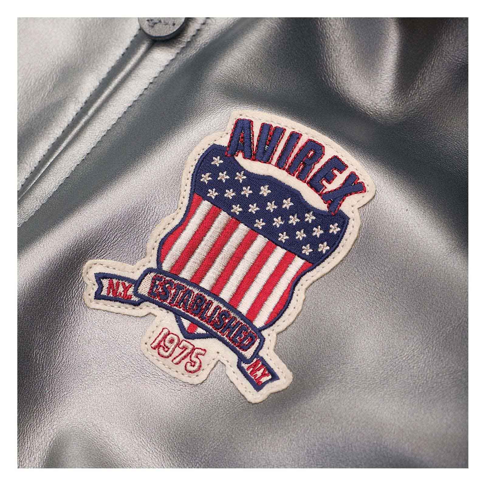 A close-up of a metallic silver leather jacket featuring a prominent embroidered patch. The patch is shaped like a shield with red and white stripes, white stars on a blue background, and a red banner reading 'AVIREX ESTABLISHED 1975 NY'.