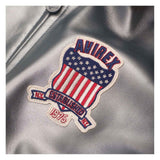 A close-up of a metallic silver leather jacket featuring a prominent embroidered patch. The patch is shaped like a shield with red and white stripes, white stars on a blue background, and a red banner reading 'AVIREX ESTABLISHED 1975 NY'.