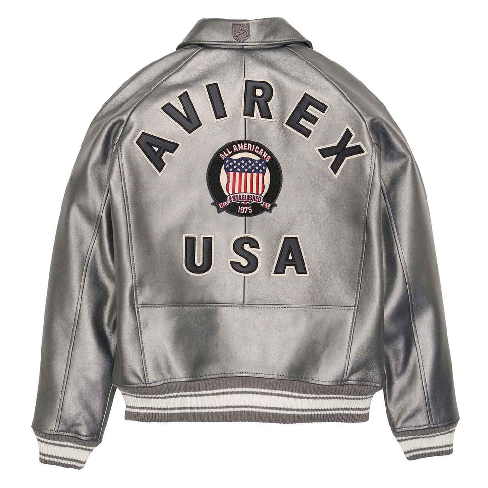 A silver metallic leather jacket shown from the back, featuring 'AVIREX USA' printed in black across the shoulders and back, with a circular American flag crest in the center. The jacket has ribbed black and white striped cuffs and hem.