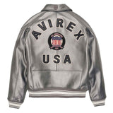 A silver metallic leather jacket shown from the back, featuring 'AVIREX USA' printed in black across the shoulders and back, with a circular American flag crest in the center. The jacket has ribbed black and white striped cuffs and hem.