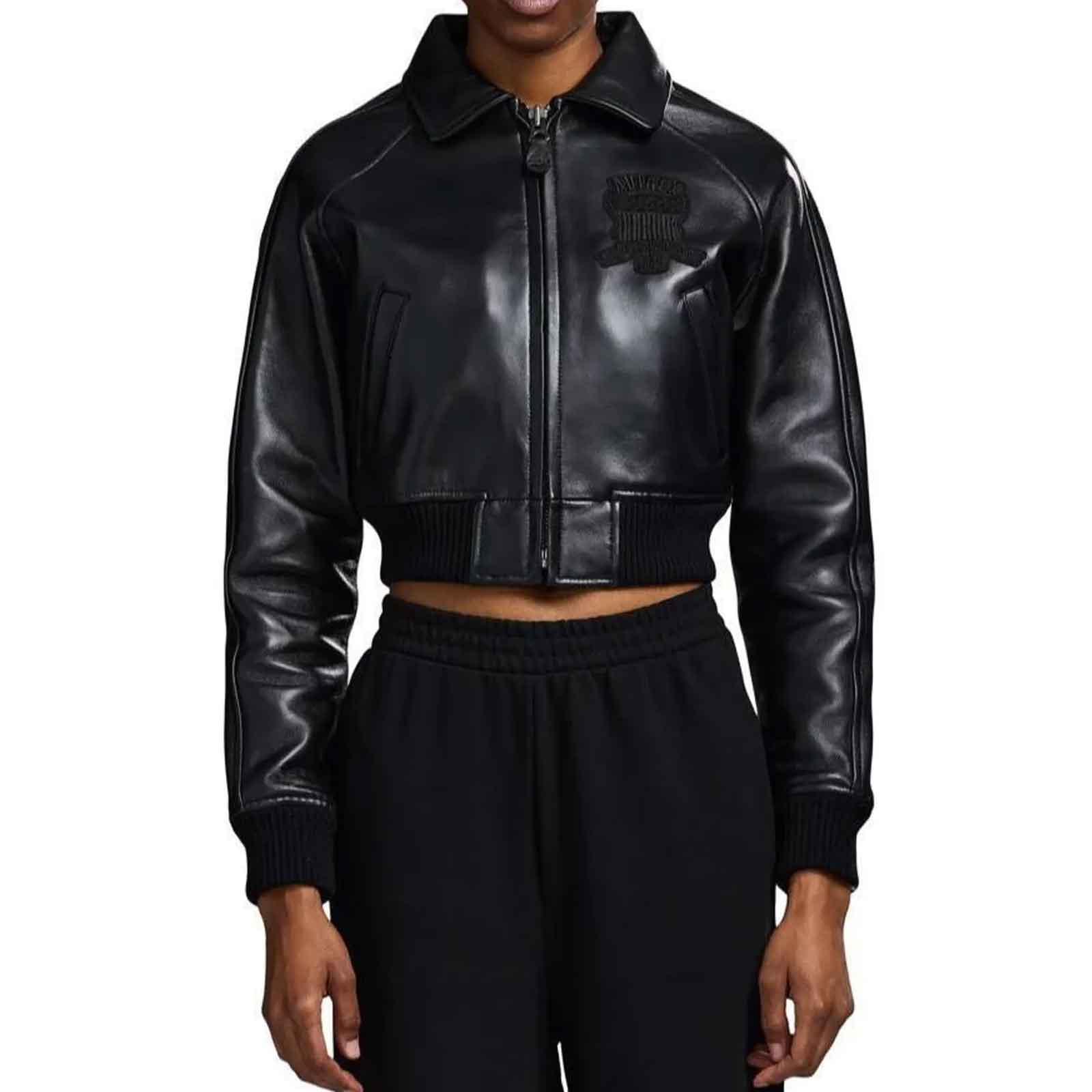 A model wearing a black faux-leather cropped zip-up jacket with a tonal black embroidered logo on the left chest, paired with black sweatpants, against a plain white background.