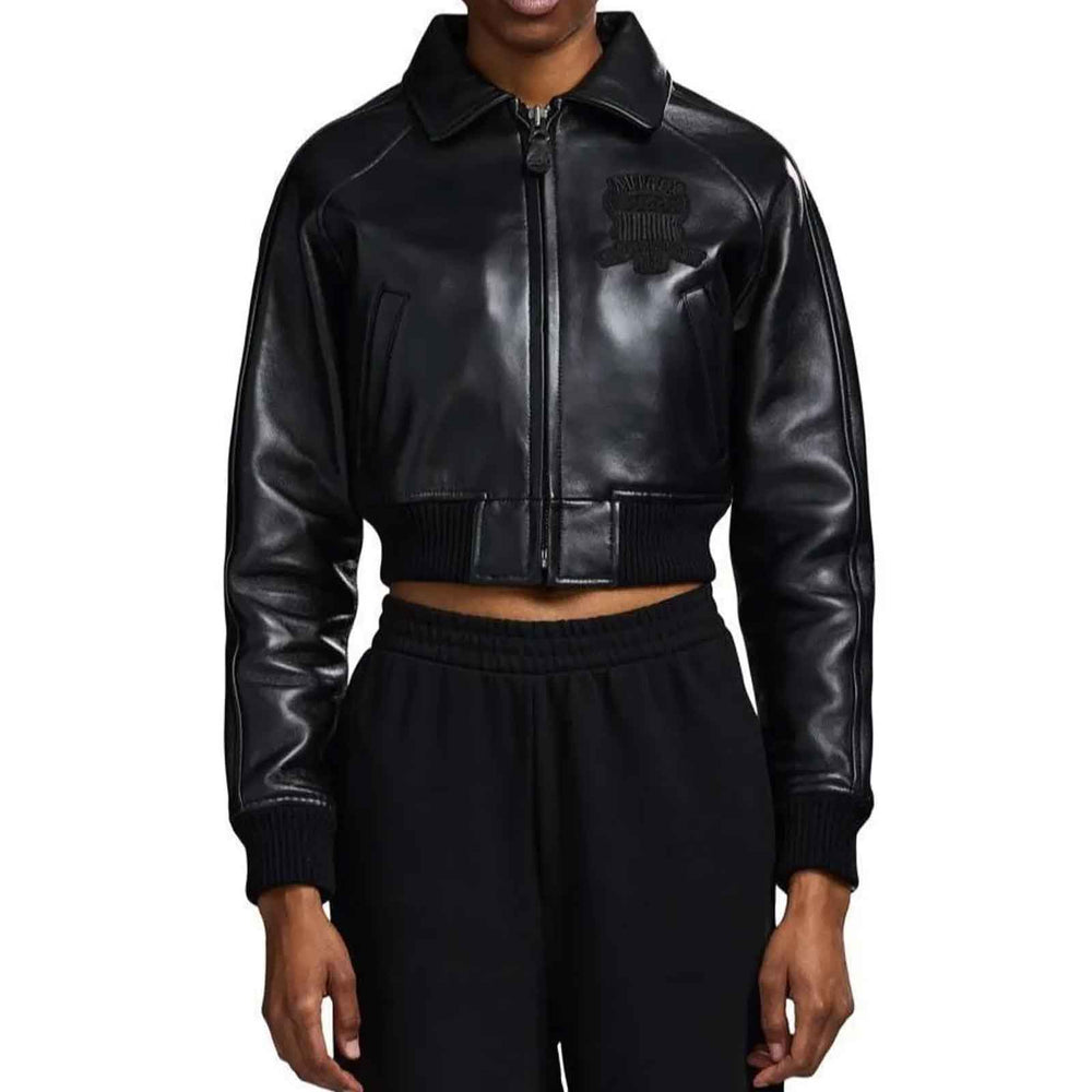 A model wearing a black faux-leather cropped zip-up jacket with a tonal black embroidered logo on the left chest, paired with black sweatpants, against a plain white background.