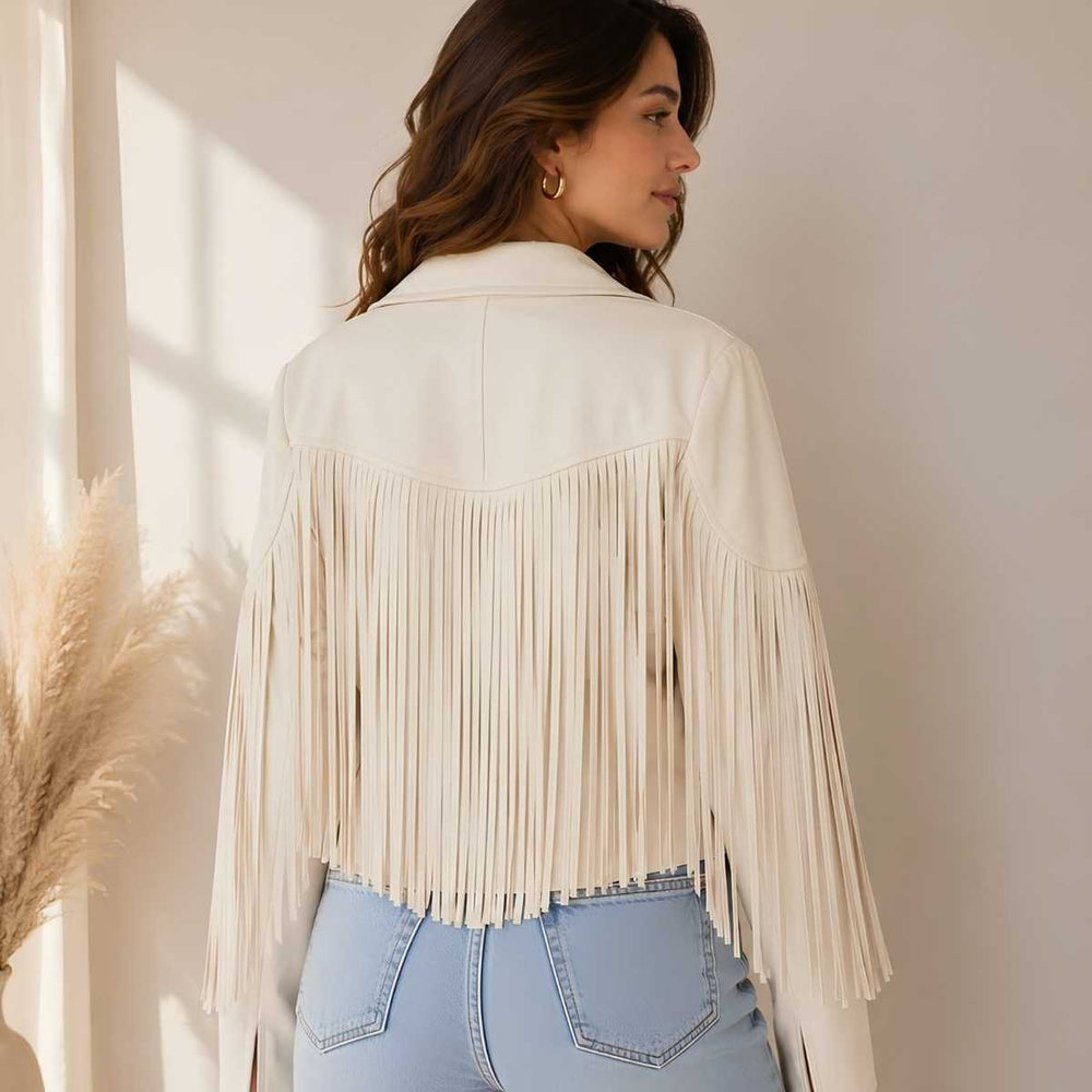 A woman shown from the back wearing a cream-colored faux leather fringe jacket over light blue jeans, standing in a brightly lit room with pampas grass on the left.