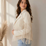 A woman wearing a cream-colored faux leather fringe jacket and light blue jeans, standing in a brightly lit room with a neutral wall and a pampas grass plant in the background.