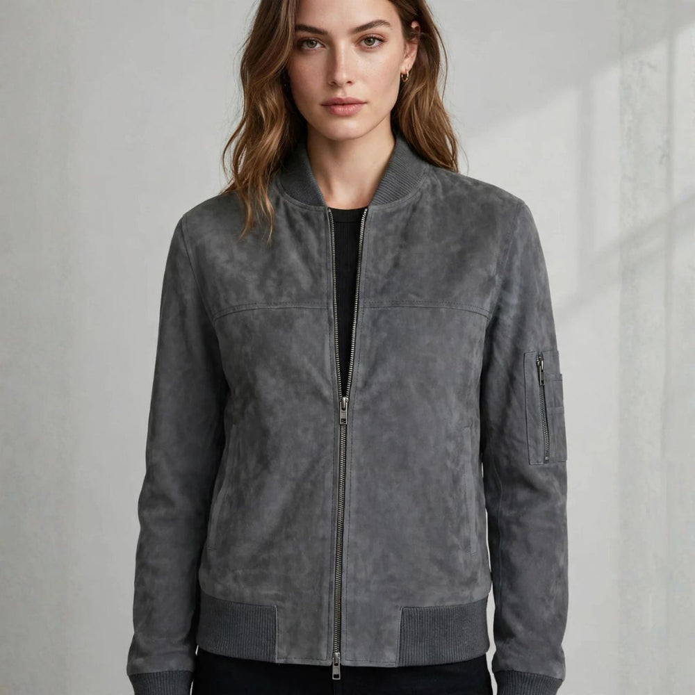 A woman wearing a gray suede bomber jacket with a front zipper, ribbed collar, cuffs, and hem, paired with a black top and black bottoms, set against a light gray textured wall.