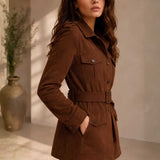 A woman wearing a brown corduroy belted jacket with chest and waist pockets, standing in a softly lit indoor setting with a large vase in the background.